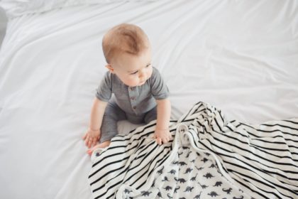 Baby in grey clothes sitting on white linen clutching dinosaur blanket